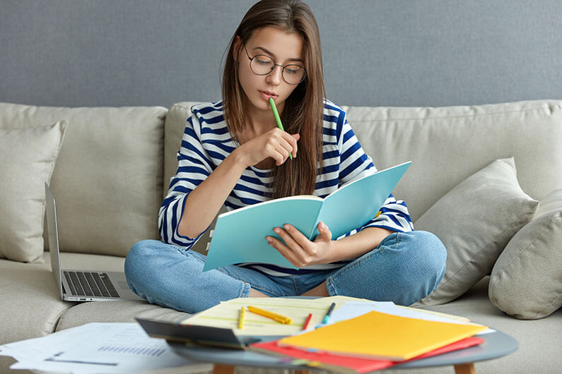 Mulher jovem séria ocupada com um projeto freelance remoto, senta-se em um sofá confortável, faz anotações, segura o livro, usa o laptop em casa com internet sem fio